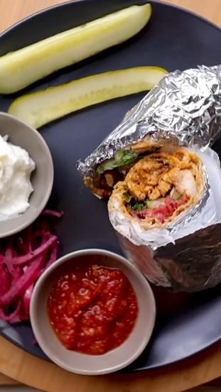 A burrito wrapped in foil next to bowls of salsa and pickles.