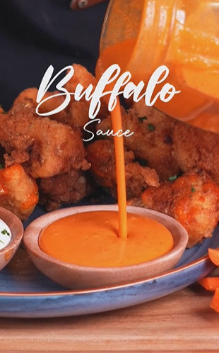 A bowl of buffalo sauce with some chicken wings in the background.