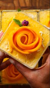 A close-up of a square serving of mango lassi tiramisu topped with a beautifully arranged mango rose, a small dried rosebud, and a fresh mint leaf. The creamy mango layer beneath has a smooth, marbled texture, and the dessert is held between two hands against a wooden background.