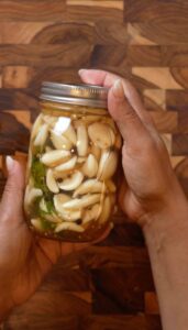 Hands holding a glass jar filled with quick pickled garlic cloves, mustard seeds, black peppercorns, and fresh dill against a wooden cutting board background.