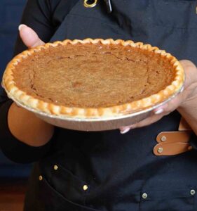 A freshly baked navy bean pie with a golden, crimped crust and smooth spiced filling, held up by a person wearing a black apron.